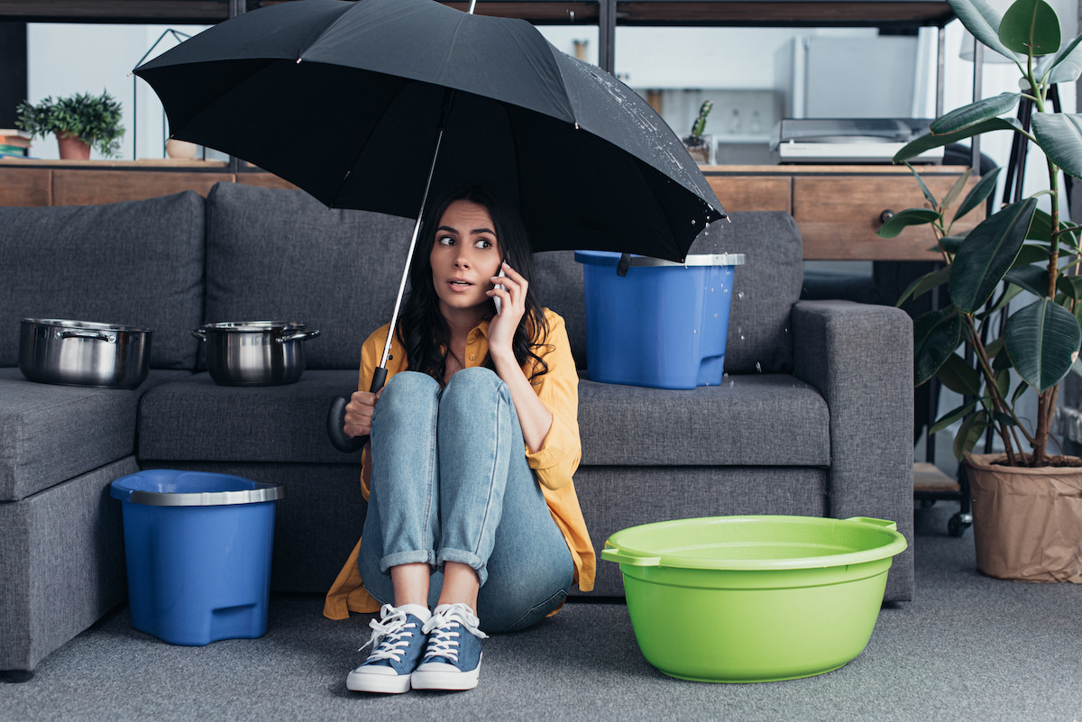 Frau sitzt mit Schirm in der Wohnung zwischen Eimern wegen eines Wasserschadens und telefoniert um Hilfe zu rufen.