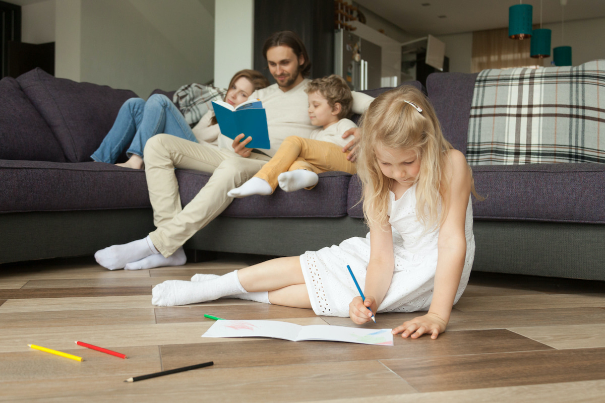 Im Vordergrund malt ein Mädchen auf dem Boden sitzend, im Hintergrund sitzen Mama, Papa und Bruder auf der Couch und Lesen.